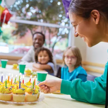 Photo shows YMCA birthday party with woman lighting cupcake candles